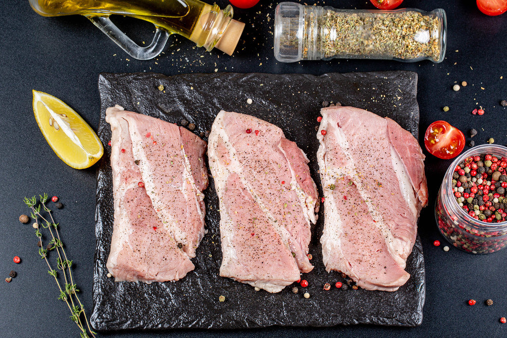 Three raw steaks with ingredients on a black background, top view