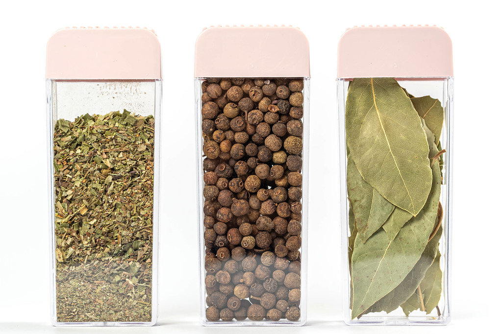 Dried Italian herbs, allspice and bay leaves in containers on white