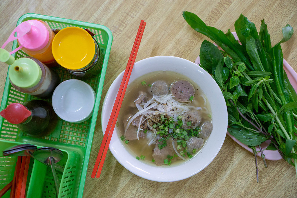 Top View Food Photo of Bowl of Vietnamse National Dish Pho Bo with Beef Balls, Fresh Herbs and Sauces on a Wooden Table