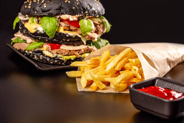 Delicious black burger with vegetables, cutlet and cheese on a dark background with fries and sauce