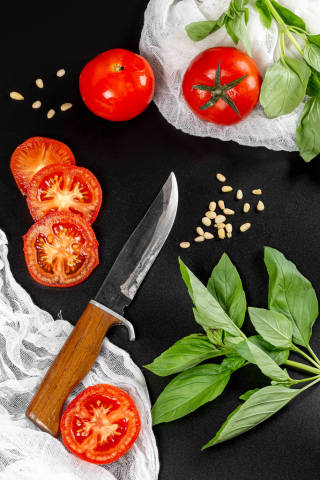 Pieces of tomatoes with basil branches, pine nuts and a knife on a dark background