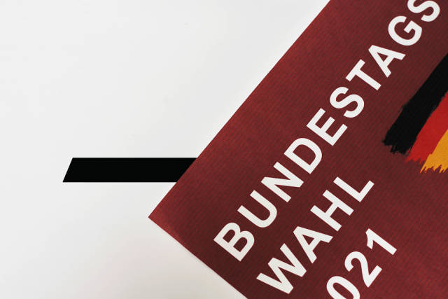 Blatt mit der Schrift "Bundestagswahl 2021" in Weiß auf Rot und der deutschen Flagge vor der Wahlurne