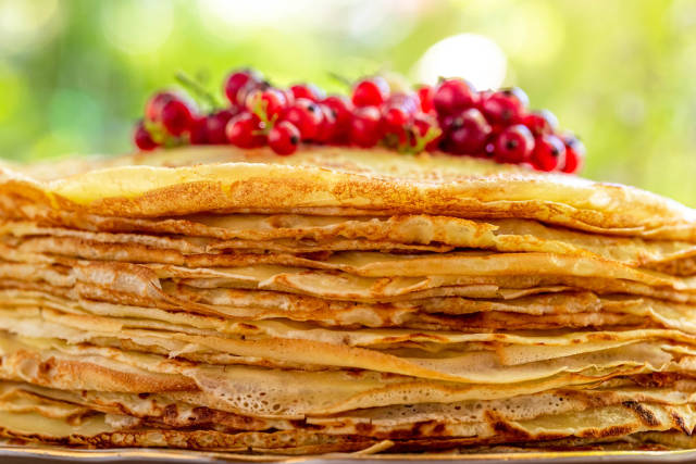 Many pancakes are stacked with fresh red currant berries on a blurred bokeh nature background