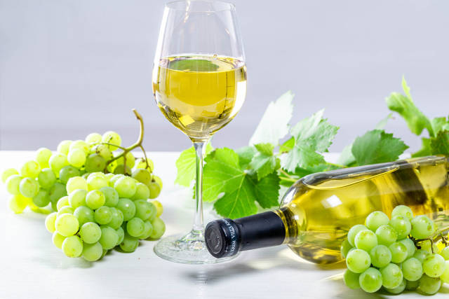 White wine in a glass with a full bottle, grapes and leaves on a white wooden background