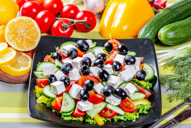 Blick von oben auf Teller mit griechischem Salat mit Oliven und Fetakäse umgeben von Gemüse