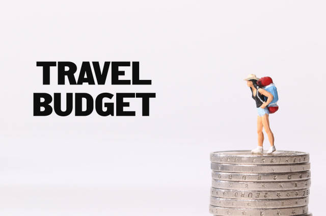 Miniature traveler standing on stack of coins and Travel Budget text
