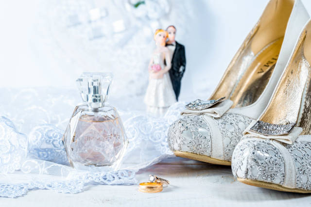Hochzeits-Szenario mit Trauringen, Parfumflakon, Brautschuhen und Brautpaar-Figur auf weißer Spitze