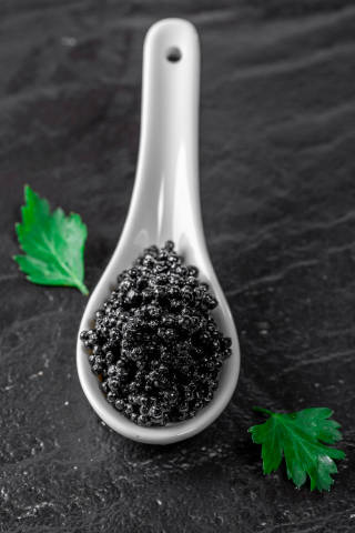 Black caviar in white ceramic spoon with parsley leaves on black background
