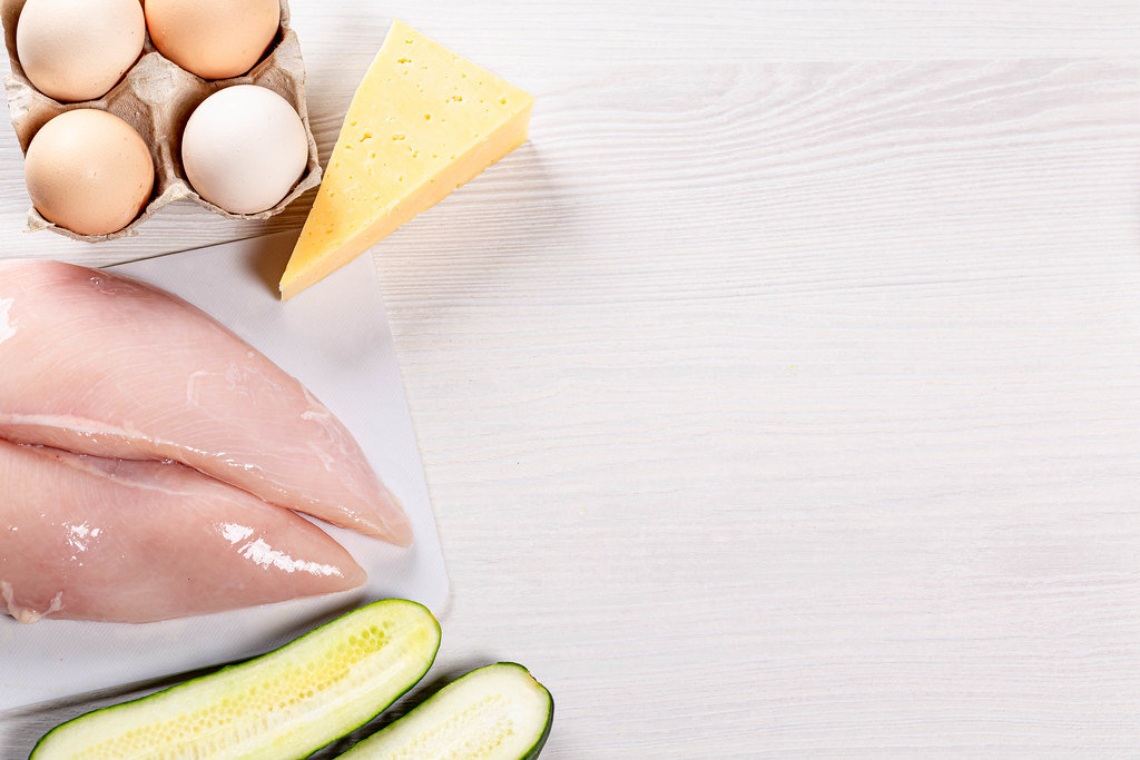 Eggs, cheese, raw chicken breast and cucumber on white wooden background