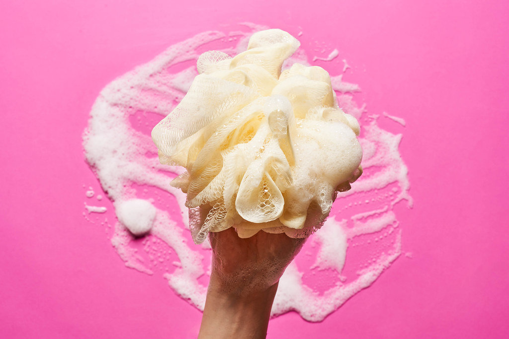 A hand of female holds skin sponge with foam over pink background