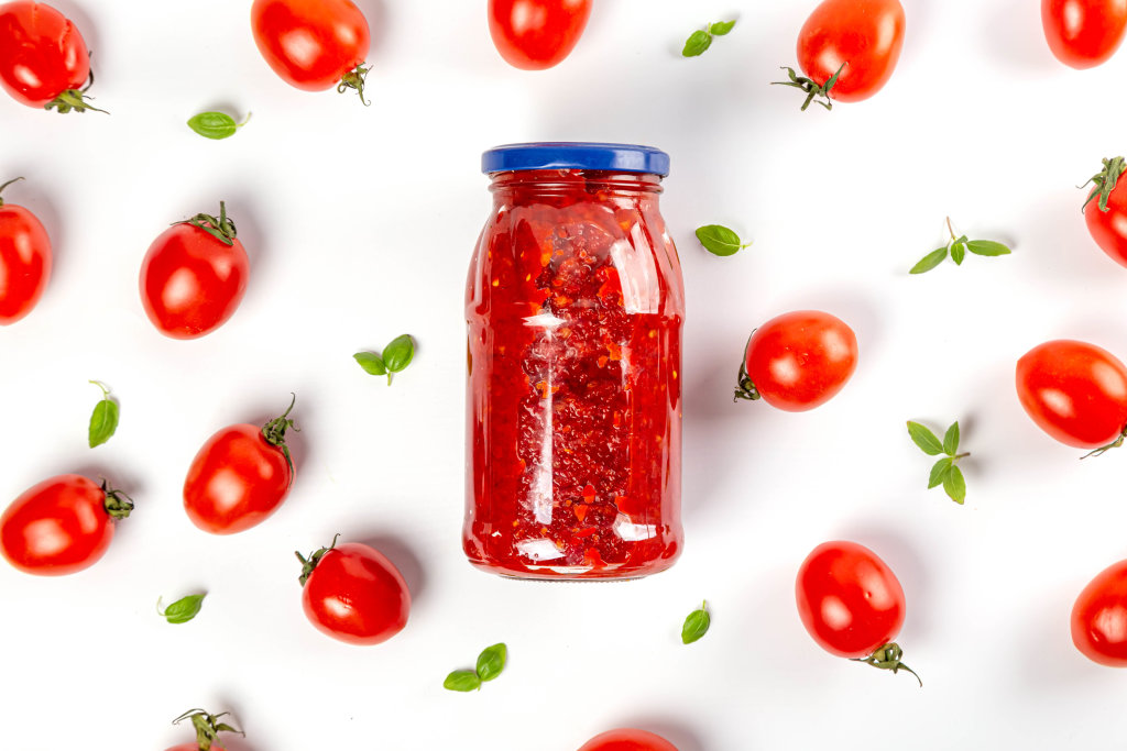 A jar of tomato sauce or ketchup on a white background with cherry tomatoes and basil leaves