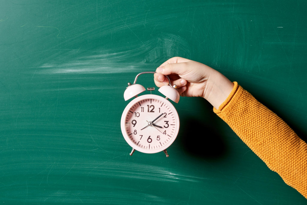 A schoolboy holds an alarm clock over the green chalkboard