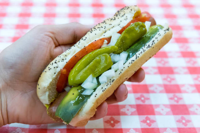 Chicago-style hot dog with yellow mustard, chopped white onions, bright green sweet pickle relish, a dill pickle spear, tomato slices and pickled sport peppers