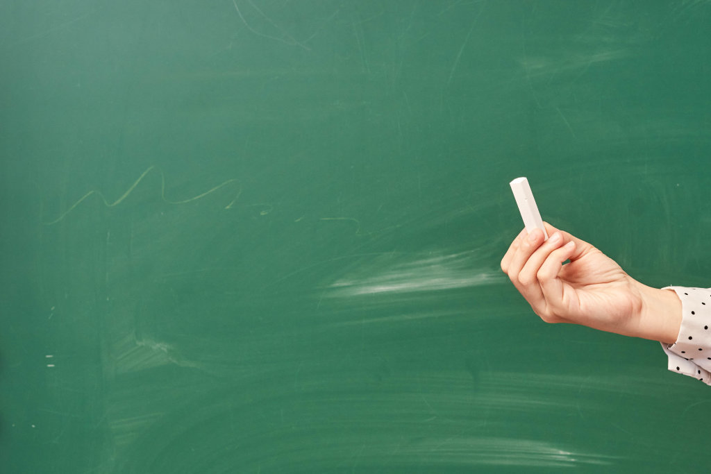 A teacher holding a piece of chalk over the green chalkboard