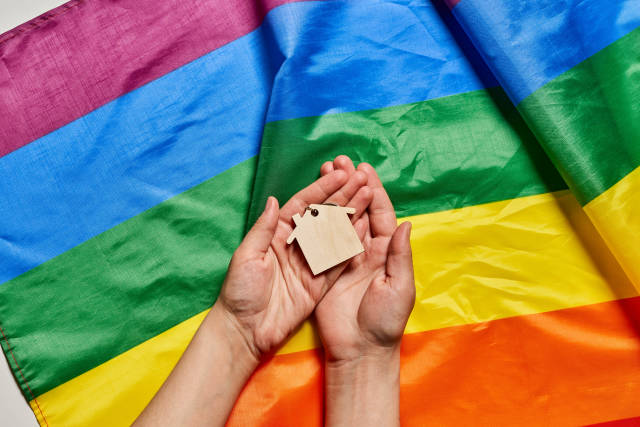 Female hand holding small wooden house over rainbow gay pride LGBT flag