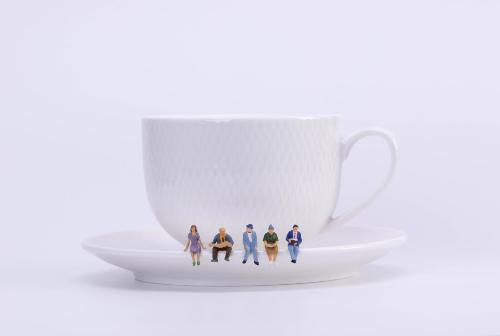 Group of miniature people figures sitting, waiting and reading newspaper and book on white plate of cup of hot coffee