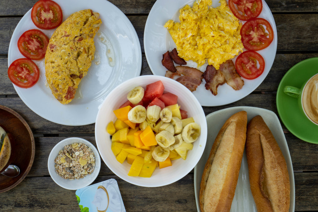 Top View Food Photo of Breakfast Table with Mixed Fruit Salad, Muesli with Yogurt, Omelette with Bacon and Tomatoes,  Scrambled Eggs with Bacon Strips, Baguettes and Coffee
