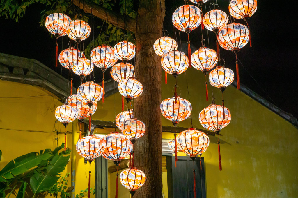 Colorful Handmade Lanterns with Light Bulbs hanging on a Tree in front of a Yellow Building in Hoi An, Vietnam