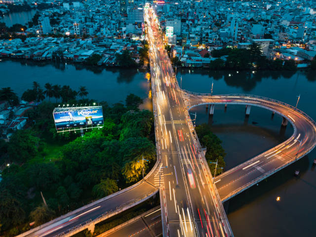 Drohnenaufnahme von einem kleinen Stadtpark, Gebäuden und Verkehr auf einer Brücke über dem Saigon Fluss mit Langzeitbelichtung in Ho Chi Minh Stadt, Vietnam