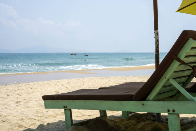 Wooden Sun Bed with Cushion and Sun Umbrella at An Bang Beach with View of Fishing Boats in Hoi An, Vietnam