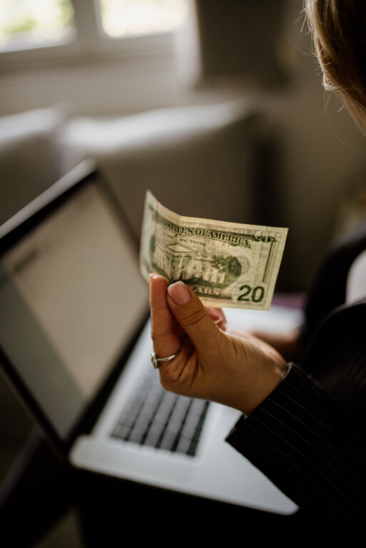 Woman holding a twenty-dollar bill while using a laptop