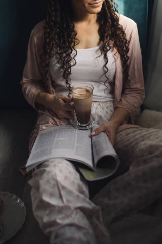 A woman in pajamas drinkig coffee and reading fashion magazine