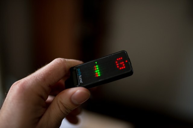 A musician holds a compact handheld tuner with green and red lights indicating pitch accuracy