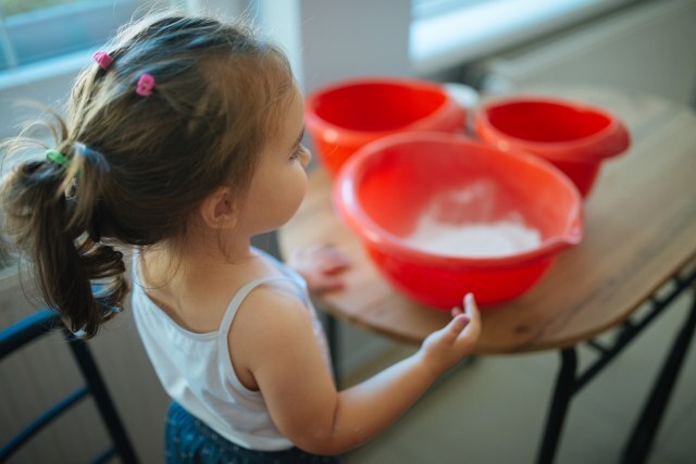 In a bright kitchen, a young girl combines ingredients into a red bowl, focused on her baking task